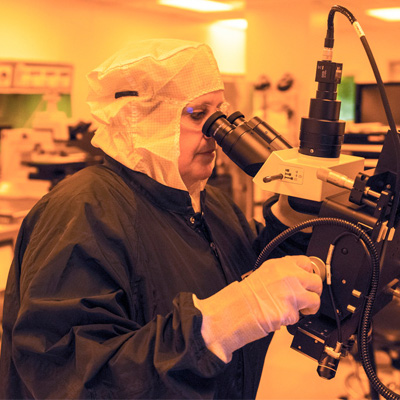 Photograph of a scientist working in a lab lit by yellow light. The scientist is wearing a protective hood, safety glasses, lab coat, and gloves, and is looking into a microscope.